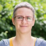 Osoba w okularach z krótkimi włosami stoi przed zielonym, rozmytym tłem i uśmiecha się delikatnie.