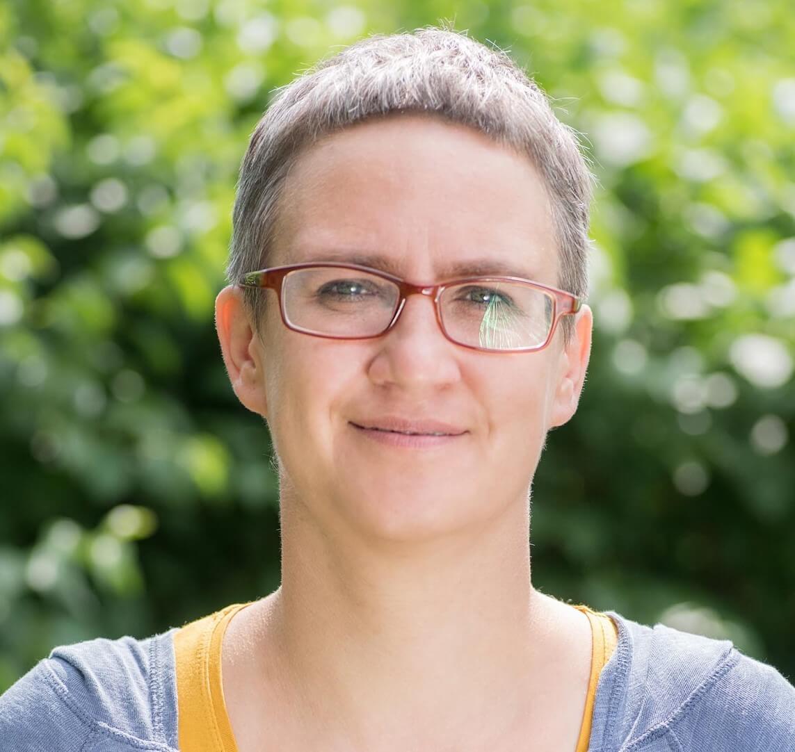 Osoba w okularach z krótkimi włosami stoi przed zielonym, rozmytym tłem i uśmiecha się delikatnie.