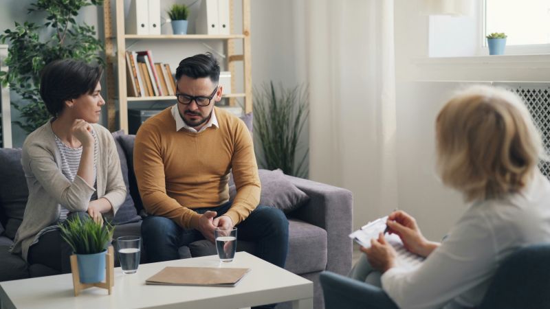 Para podczas sesji terapeutycznej z terapeutką przy stoliku, w otoczeniu roślin i książek.