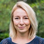 Portret uśmiechniętej kobiety z blond włosami na tle zielonej roślinności.