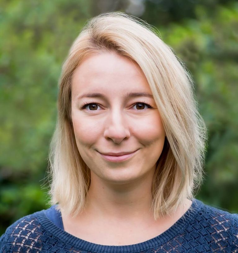 Portret uśmiechniętej kobiety z blond włosami na tle zielonej roślinności.