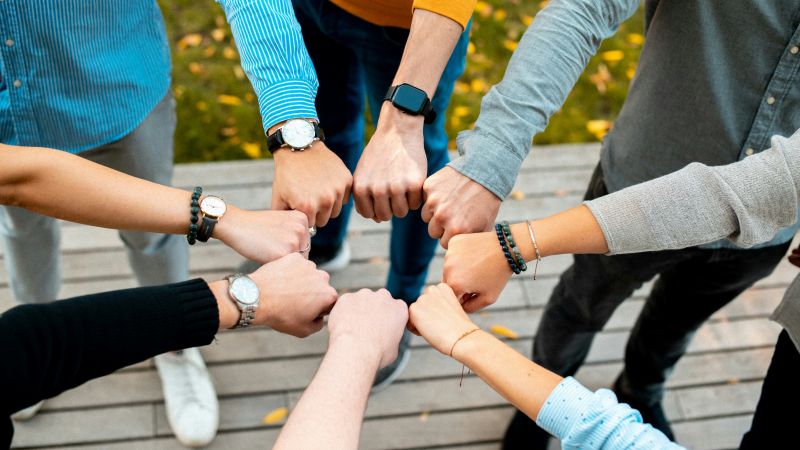 Grupa ludzi robi gest złożonymi pięściami, stojąc w kręgu na drewnianym tarasie.