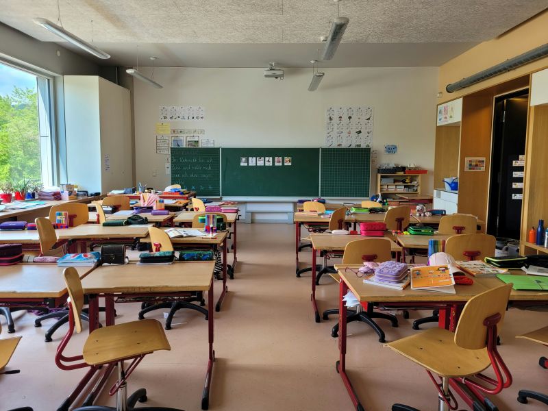 Pusta sala lekcyjna z biurkami i krzesłami, na tablicy wiszą plakaty edukacyjne, a okno wpuszcza światło słoneczne.