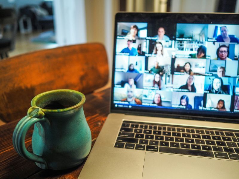Laptop z wideokonferencją na ekranie i zielony kubek z kawą na drewnianym stole.